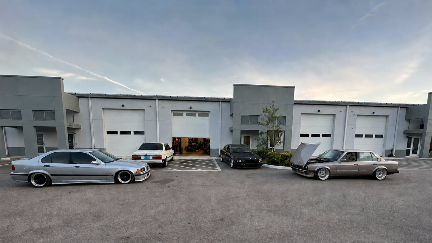 Four classic BMWs parked outside the first shop at dusk, the bay door open behind them